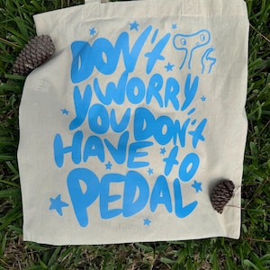 May include: A white canvas tote bag with blue text that reads "Don't worry you don't have to pedal" with a small blue alien illustration and stars.