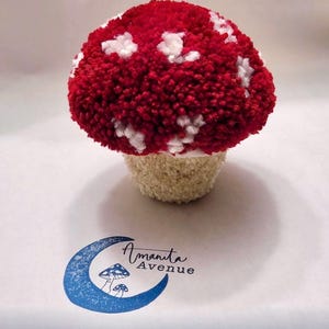 May include: A red and white yarn mushroom with white polka dots. The mushroom is made with pom-poms and is sitting on a white surface. The image also includes a blue and white logo with the text "Amanita Avenue".