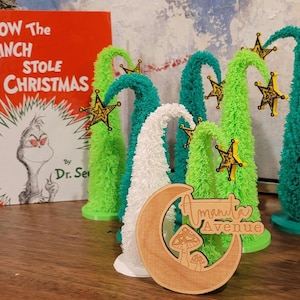 Könnte beinhalten: Ein Set aus fünf Miniatur-Weihnachtsbäumen, die mit goldenen Sternen verziert sind. Die Bäume bestehen aus grünem und weißem, flauschigem Material. Die Bäume sind in einem Halbkreis auf einer Holzoberfläche angeordnet. Ein Holzschild mit dem Text "Amanita Avenue" befindet sich im Vordergrund.