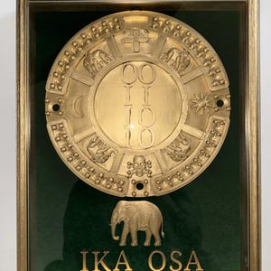 May include: A framed gold-colored relief sculpture with a circular design, featuring elephants, a cross, and the numbers "0010". Below the sculpture is a gold elephant figure and the text "IKA OSA". The background is dark green.