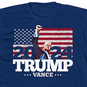 May include: Navy blue t-shirt featuring a graphic of a man raising his fist in front of an American flag. The text "2024 TRUMP VANCE" is displayed in white, with stars below the names. The design has a distressed, vintage look.