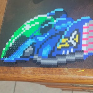 May include: A blue, green, white, and gray pixel art creation of a race car. The car is made of small plastic beads.