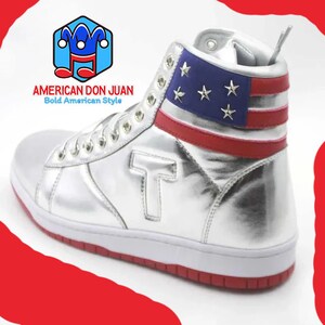 Puede incluir: Zapatillas de deporte de ca&ntilde;a alta plateadas con un dise&ntilde;o de bandera estadounidense rojo, blanco y azul en la parte superior del zapato. El zapato tiene una suela blanca con un ribete rojo.