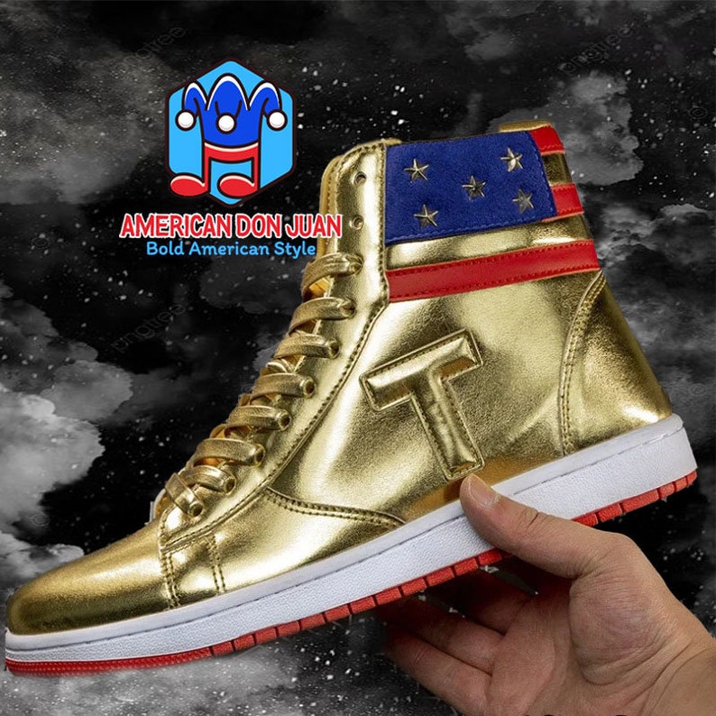 Puede incluir: Zapatillas doradas de ca&ntilde;a alta con un dise&ntilde;o de bandera americana roja y azul en la parte posterior. Las zapatillas tienen una suela blanca y una franja roja alrededor de la parte inferior. Las zapatillas tienen una gran letra "T" dorada en el lateral. El texto "AMERICAN DON JUAN Bold'American Style" est&aacute; en la imagen.
