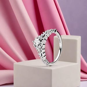 Può includere: Un anello d'argento con un design a corona, con una grande gemma rotonda e trasparente al centro e pietre più piccole lungo la fascia. L'anello è esposto su un blocco bianco su uno sfondo rosa e viola.