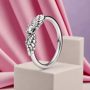 Op de afbeelding: Een zilveren ring met een gedetailleerd vleugelontwerp, met een centrale heldere edelsteen en kleinere accentstenen. De ring wordt gepresenteerd op een witte ondergrond tegen een roze en paarse achtergrond, wat de elegante stijl benadrukt.