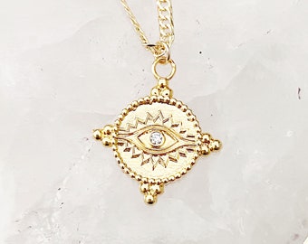 May include: Gold necklace with a small, round pendant featuring a detailed eye design with a single clear stone in the centre. The pendant is surrounded by a beaded border.