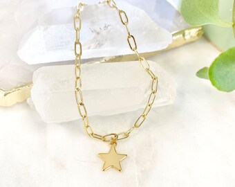 May include: Gold chain bracelet with a star pendant. The bracelet has rectangular links and a five-pointed star charm. The jewellery is displayed on a white surface with a crystal and greenery in the background.