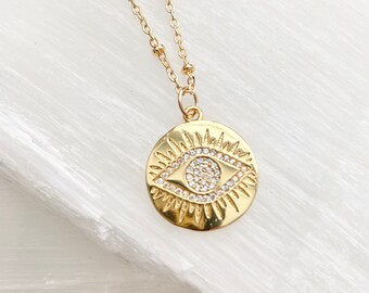 May include: Gold necklace with a round pendant featuring a detailed evil eye design with clear sparkling stones.