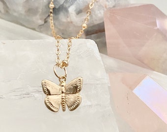 Pode incluir: Um colar de corrente delicada com um pingente de borboleta de ouro. O colar é exibido em uma superfície branca com cristais rosa e iridescentes.