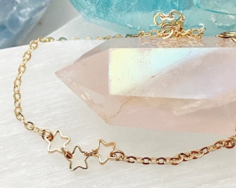 May include: A gold chain bracelet with three star charms. The bracelet is lying on a white surface with a pink crystal next to it.