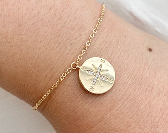 May include: A gold-colored bracelet featuring a round pendant with a compass design. The pendant has a starburst pattern of small, clear stones. The bracelet is on a delicate chain.