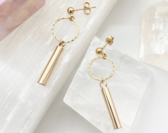 May include: Gold earrings with a small circle and a dangling cylinder. The earrings are on a white surface.