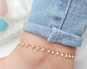 May include: A gold chain anklet with small white beads dangling from it. The anklet is worn on a person's ankle.