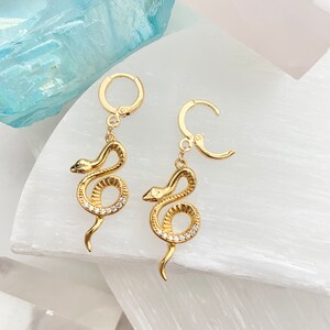 May include: A pair of gold snake earrings with clear crystal accents. The earrings have a hoop design and dangle from the hoop.