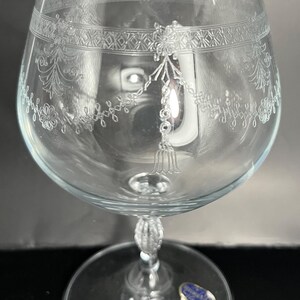 May include: A clear glass brandy snifter with an etched design around the bowl. The snifter has a round base and a stem with a decorative knob. The glass has a blue sticker on the base that reads "Made in Czechoslovakia".