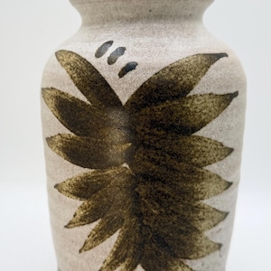 May include: A ceramic vase with a beige background and a dark brown floral design. The vase has a cylindrical shape with a slightly flared rim and a dark brown base. The floral design is composed of stylized leaves.
