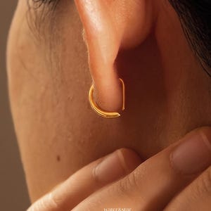 May include: A small, polished gold hoop earring with a smooth, rounded surface and a minimalist open-ended design. The earring is worn in the earlobe, featuring a curved, continuous metal band that creates a simple, elegant circular silhouette.