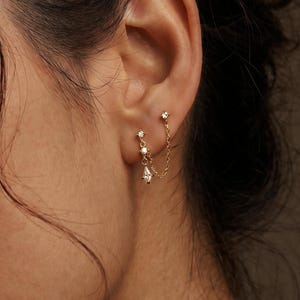 May include: A pair of gold earrings with a delicate chain. One earring features a small stud with a single gem, connected by a chain to a second earring with a teardrop-shaped gem and two smaller gems.