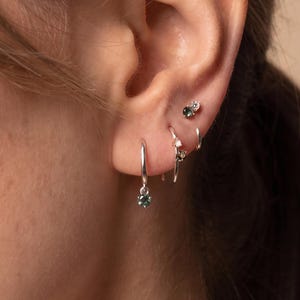 May include: A set of silver earrings worn in the ear, featuring a small hoop with a dangling dark green gemstone, a thin silver hoop, and a stud earring with a small dark green gemstone and a tiny clear accent stone.