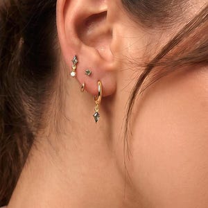 May include: A close-up of a person's ear adorned with multiple gold earrings. The earrings include small studs, a hoop earring with a dangling charm, and a delicate earring with a small pearl. The jewelry is set against a neutral background.