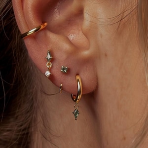 May include: A gold ear cuff, a small gold hoop earring with a dangling green marquise-cut stone and a clear round stone, a tiny green stud earring, and a gold hoop earring with a dangling green marquise-cut stone worn in a single ear.