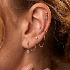 Puede incluir: Juego de aretes dorados con múltiples piezas. Incluye un arete de botón con una gema colgante en forma de lágrima, una cadena que conecta dos aretes y un arete de puño. Los aretes tienen pequeñas gemas transparentes.