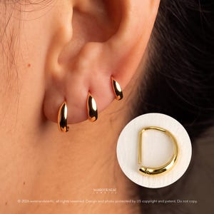 May include: Three small, polished gold hoop earrings worn in a row along the earlobe. A circular inset shows a single gold hoop earring with a smooth, curved design and a small, subtle star-shaped engraving near the closure point.