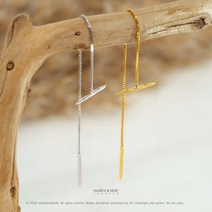 May include: A pair of delicate threader earrings, one silver-toned and the other gold-toned, each featuring a linear chain and a small, stylized wing-shaped charm. The earrings are displayed against a natural wood branch, with the brand name "water&star jewelry" visible.