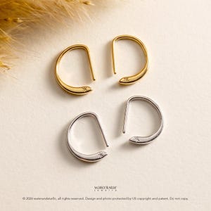Pode incluir: Dois pares de brincos em forma de U, um par em tom dourado e outro em tom prateado. Cada brinco apresenta um design curvo e liso com um pequeno detalhe de estrela gravado na extremidade. O texto water&star jewelry é visível abaixo.