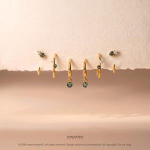 May include: A set of gold hoop earrings featuring small, round, dark green gemstones and clear accent stones. The jewelry pieces are arranged in a row, showcasing delicate metal bands and secure clasps, with the text water&star jewelry visible.