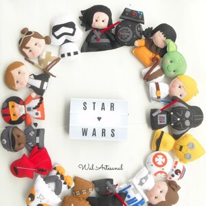 Puede incluir: Un círculo de personajes de Star Wars de fieltro, incluyendo Darth Vader, Yoda, R2-D2 y Chewbacca. Los personajes están dispuestos alrededor de una caja de luz con las palabras "STAR WARS" en ella.