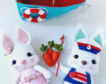 May include: Two white felt bunny plushies, one wearing a pink dress and the other a blue sailor outfit with a red anchor. The bunnies are holding a red felt carrot and a wooden stick. A blue and red felt sailboat with a white sail is in the background.