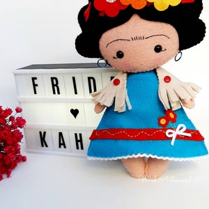 May include: A felt doll with black hair, a blue dress, and a white shawl. The doll is wearing a flower crown with red, orange, and yellow flowers. The doll is sitting in front of a white light box with the word "FRIDAH" spelled out in black letters.