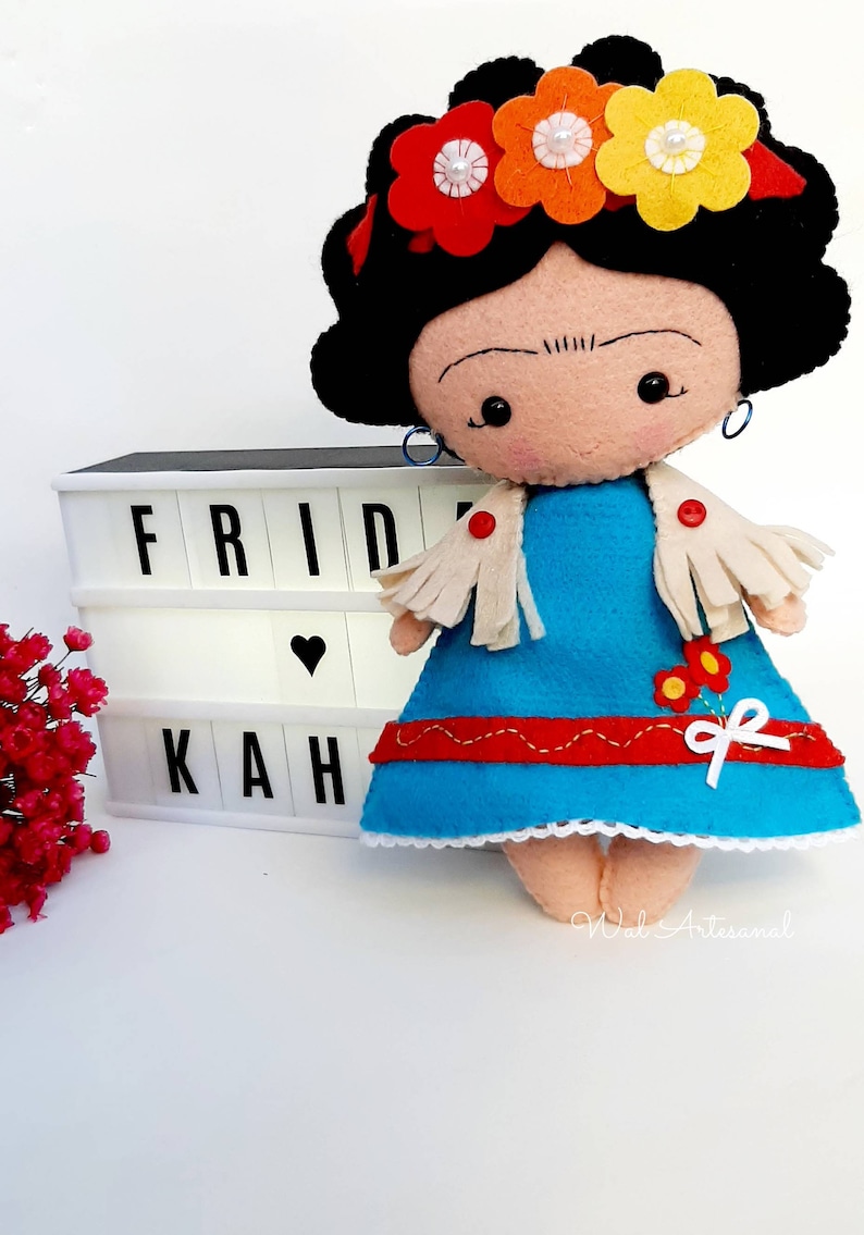 May include: A felt doll with black hair, a blue dress, and a white shawl. The doll is wearing a flower crown with red, orange, and yellow flowers. The doll is sitting in front of a white light box with the word "FRIDAH" spelled out in black letters.