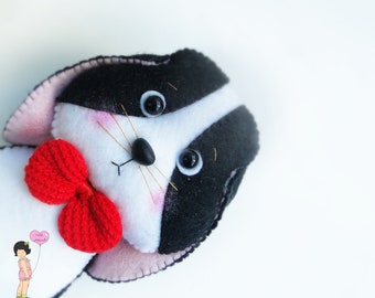 May include: A black and white plush toy with a red bow tie. The toy has a white face with black eyes and a black nose. The toy is made of felt and has a soft, cuddly texture.