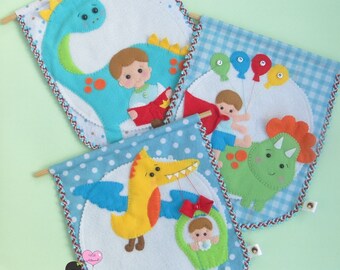May include: Three handmade felt banners with dinosaur and baby boy designs. Each banner features a different dinosaur: a blue Brontosaurus, a green Triceratops, and a yellow Pterodactyl. The banners have blue polka dot and gingham backgrounds.