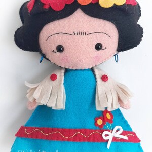May include: A felt doll with black hair, a blue dress, a white shawl, and a flower crown. The doll has a painted face with black eyes and a small smile. The doll is wearing red buttons on the shawl and has a white bow on the dress. The doll is handmade and has the text "Wal Artesanal" on the bottom of the dress.