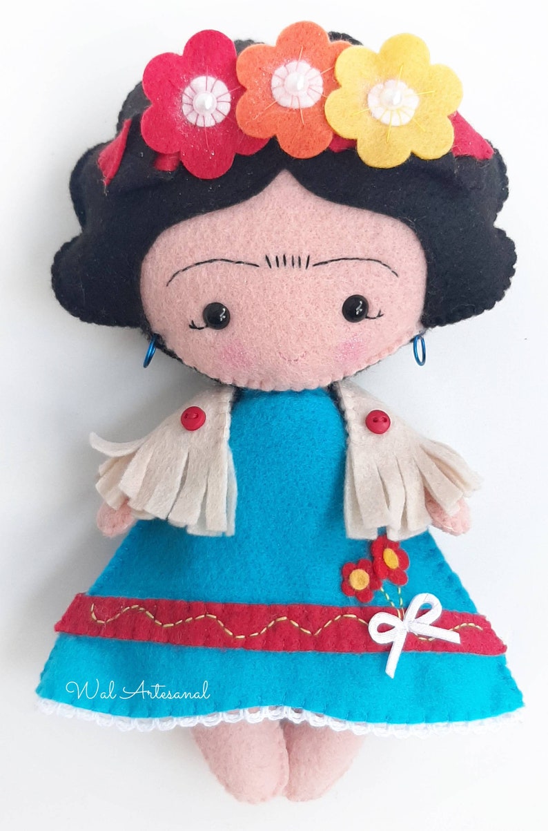 May include: A felt doll with black hair, a blue dress, a white shawl, and a flower crown. The doll has a painted face with black eyes and a small smile. The doll is wearing red buttons on the shawl and has a white bow on the dress. The doll is handmade and has the text "Wal Artesanal" on the bottom of the dress.