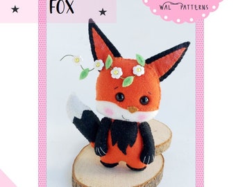 May include: A felt fox toy with a white flower crown, orange body, black legs and tail, and a white tip on the tail. The toy is sitting on a wooden slice. The image includes the text "FOX" and "WAL PATTERNS".