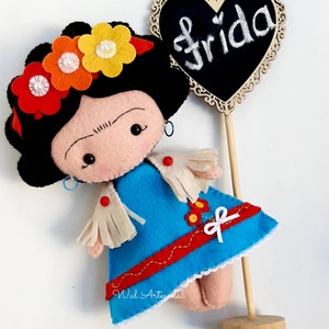 May include: A felt doll of Frida Kahlo with a black flower crown, blue dress, and white fringe. The doll is holding a wooden sign with the word "Frida" written on it in white chalk.