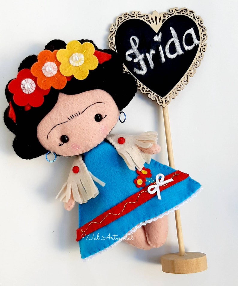 May include: A felt doll of Frida Kahlo with a black flower crown, blue dress, and white fringe. The doll is holding a wooden sign with the word "Frida" written on it in white chalk.