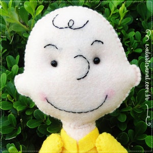 Op de afbeelding: Een wit pluche speelgoed van vilt met een geel shirt. Het speelgoed heeft een zwart geborduurd gezicht met een glimlach en zwarte ogen. Het speelgoed is een personage uit de strip Peanuts.