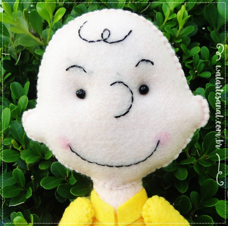 Op de afbeelding: Een wit pluche speelgoed van vilt met een geel shirt. Het speelgoed heeft een zwart geborduurd gezicht met een glimlach en zwarte ogen. Het speelgoed is een personage uit de strip Peanuts.