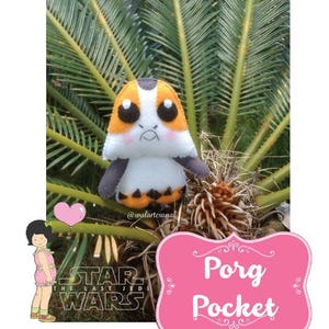 May include: A plush toy of a Porg from Star Wars: The Last Jedi. The Porg is orange, white, and black with a cute expression. The toy is sitting on a green plant. The text "Porg Pocket" is on a pink sign.