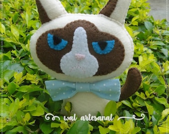 May include: A plush toy cat with brown fur, white facial markings, and blue eyes. The cat is wearing a blue bow tie with white polka dots. The cat is sitting in a bed of green leaves.