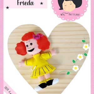 May include: A felt doll named Frieda, with red hair, a yellow dress, and black shoes, is displayed on a wooden heart-shaped plaque. The plaque has a floral design with white flowers and green leaves. The text "2024 by Wal Vieira" and "NOTHING IS SMALL IF IT'S DONE FOR LOVE" are also on the plaque.