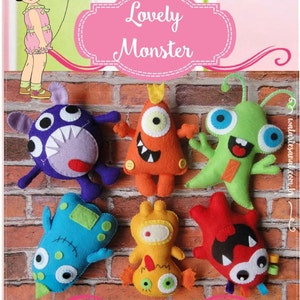 Puede incluir: Seis juguetes de monstruos de fieltro con diferentes colores, formas y expresiones. Los monstruos están dispuestos sobre un fondo de pared de ladrillo. El texto "Lovely Monster" se muestra en una pancarta rosa sobre los juguetes.