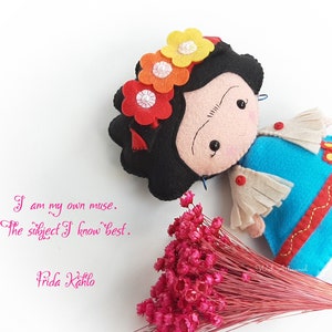 May include: A felt doll of Frida Kahlo with a black wig, a colorful flower crown, and a blue and white dress. The doll is holding a bouquet of pink flowers. The quote "I am my own muse. The subject I know best. Frida Kahlo" is written below the doll.