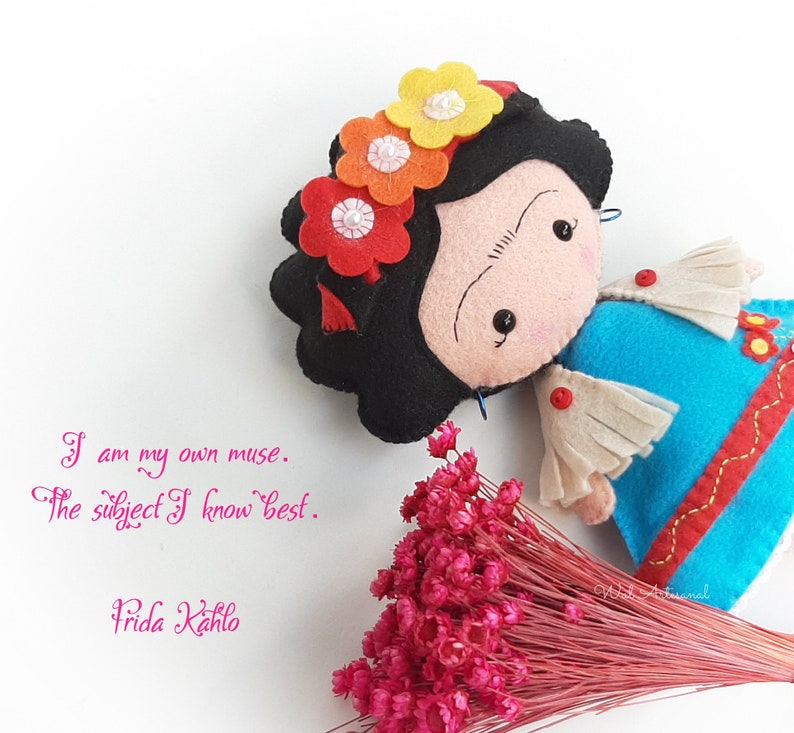 May include: A felt doll of Frida Kahlo with a black wig, a colorful flower crown, and a blue and white dress. The doll is holding a bouquet of pink flowers. The quote "I am my own muse. The subject I know best. Frida Kahlo" is written below the doll.